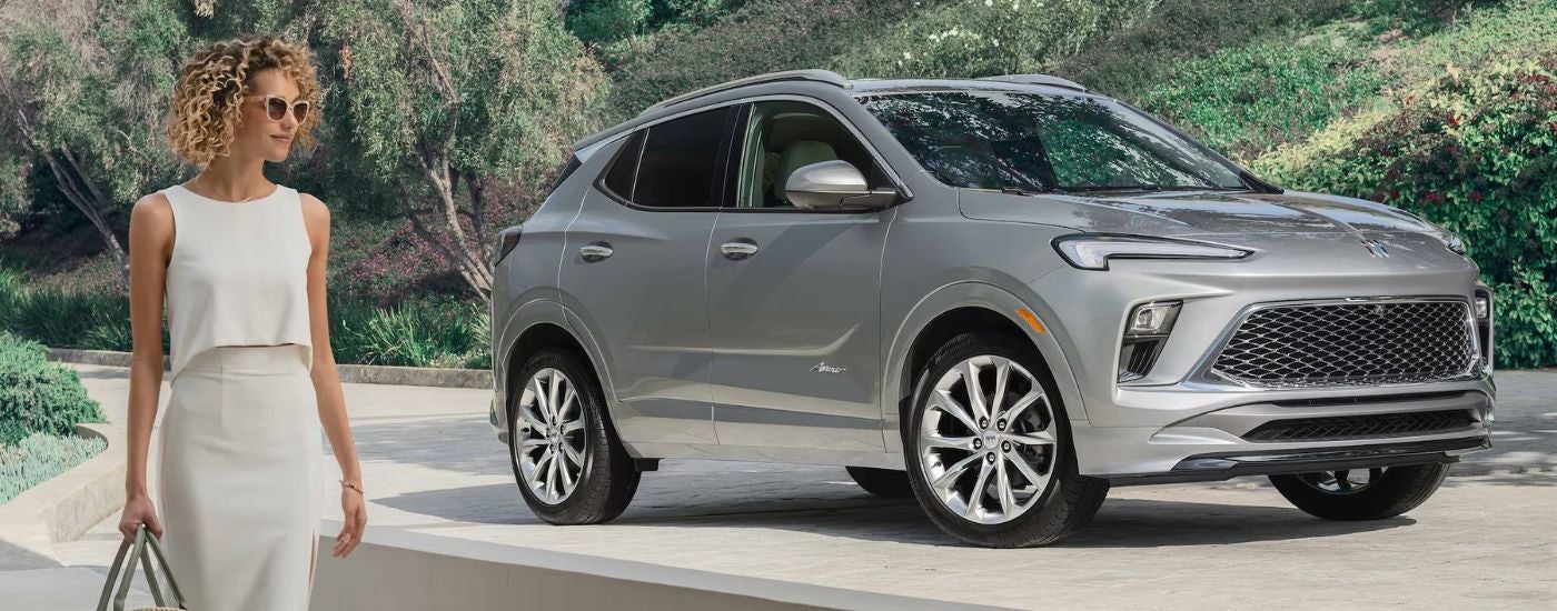 A woman walking past a silver 2025 Buick Encore GX parked on an elevated driveway.